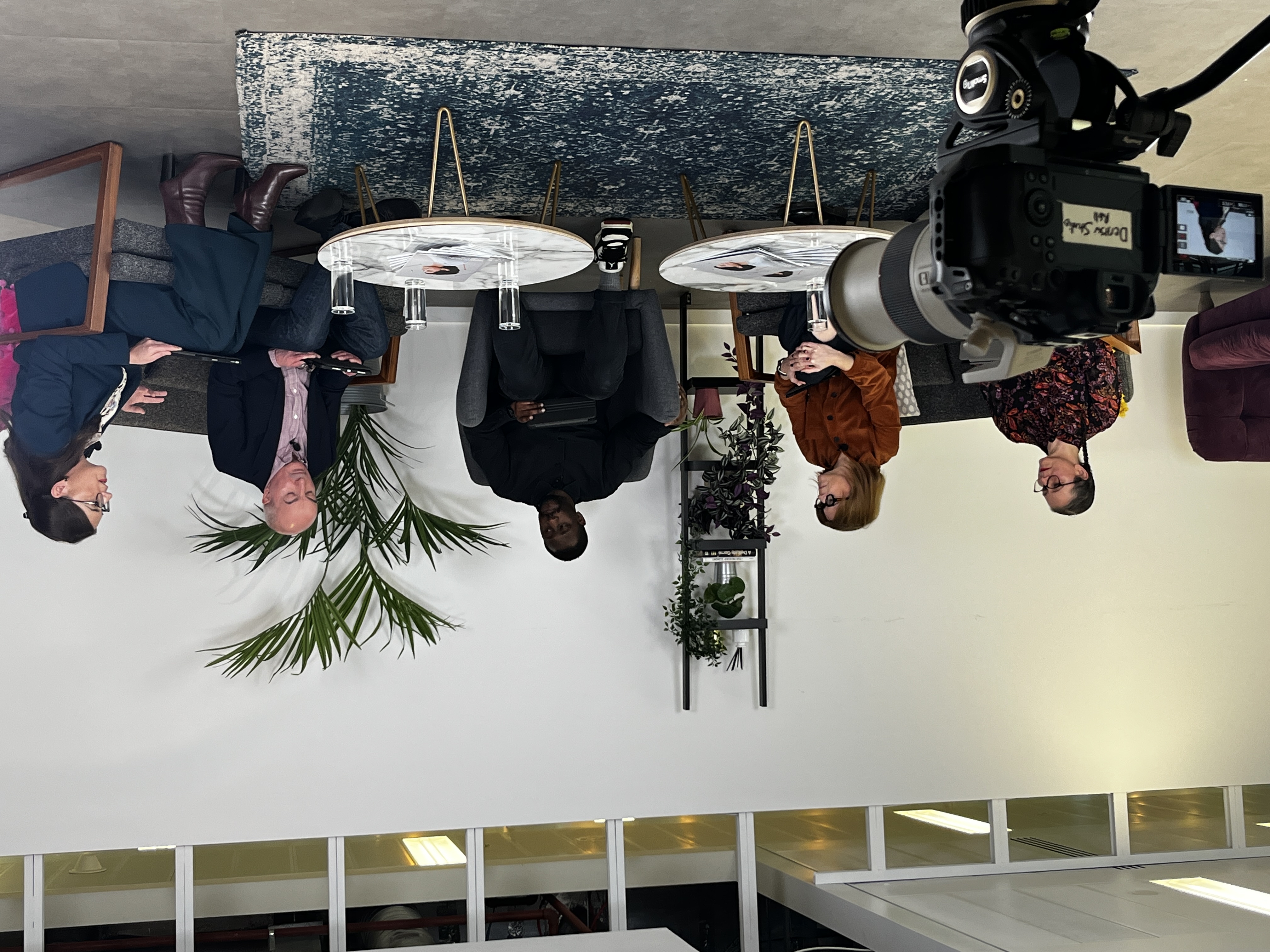 A group of five people standing together indoors, smiling at the camera. Two individuals on the left and one on the right are holding printed copies of a report featuring a person wearing headphones on the cover. They are dressed in smart and semi-formal clothing, standing in a modern studio-style setting with a rug, chairs, and soft lighting in the background.